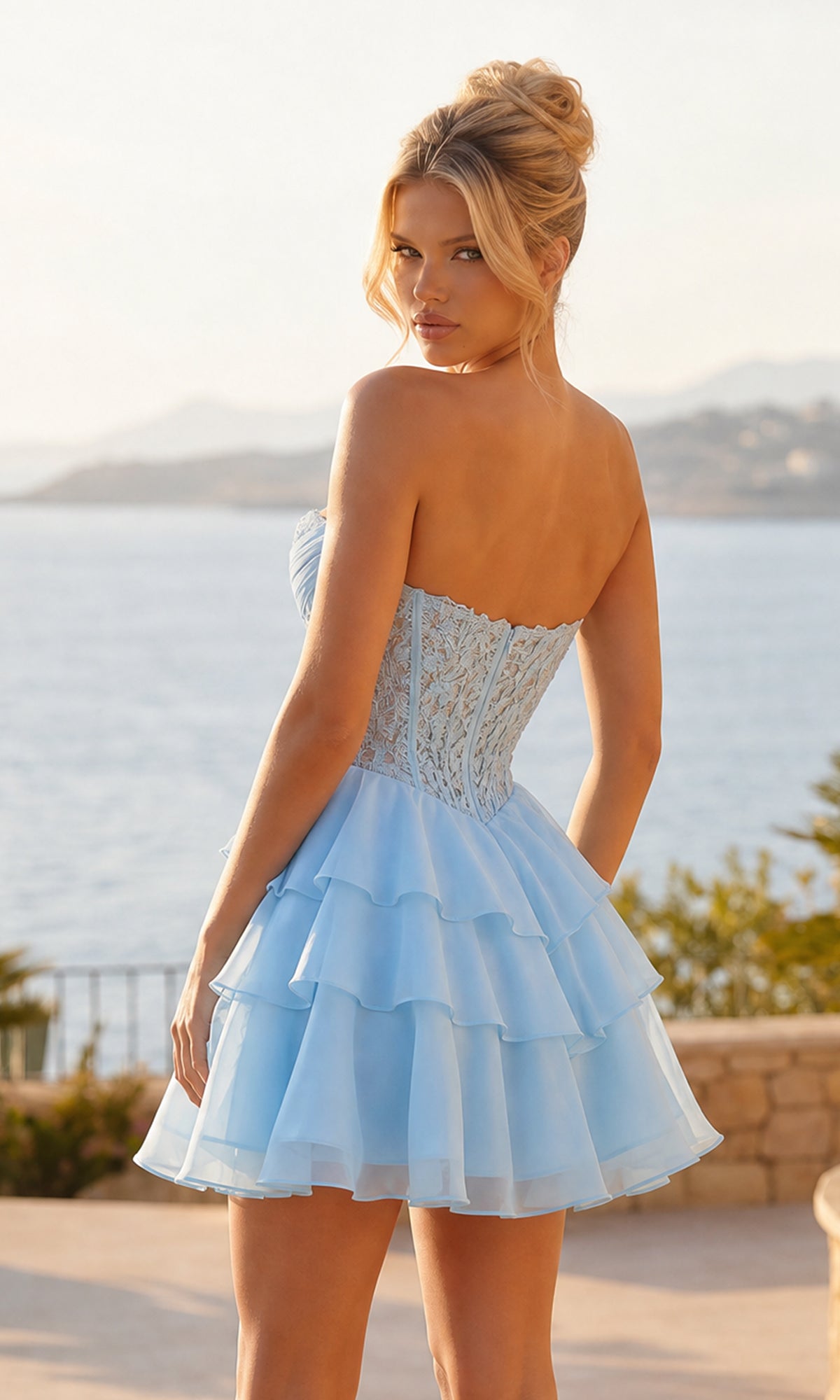 A woman stands outdoors by the water and mountains, wearing the PromGirl Exclusive 250334 short homecoming dress with a strapless bodice and light blue layered ruffle skirt.